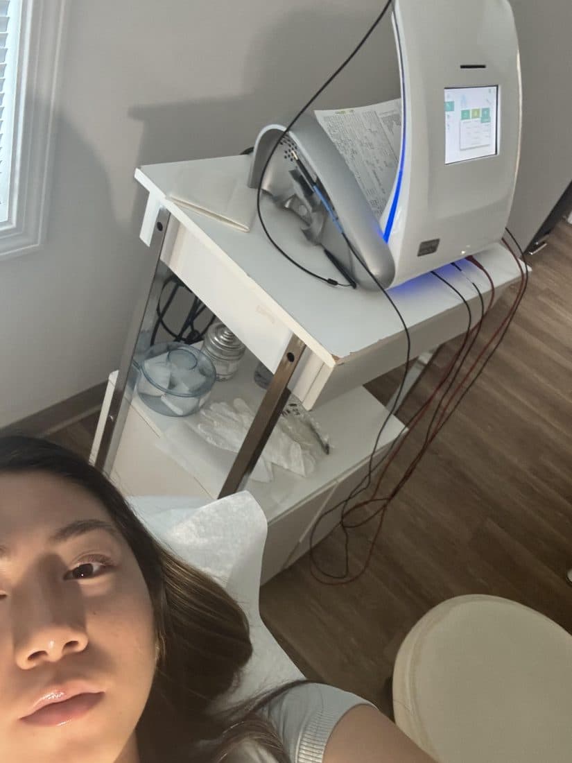 Hanna in the treatment chair as an electrolysis patient, next to the clinic's machine and instrument cart.